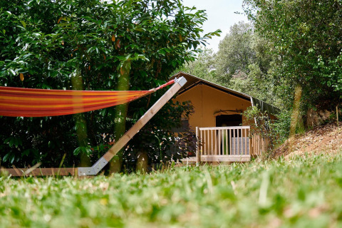 Tienda safari Villatent Wood con hamaca y terraza, rodeada de vegetación en Camping Le Pech Charmant, Francia.