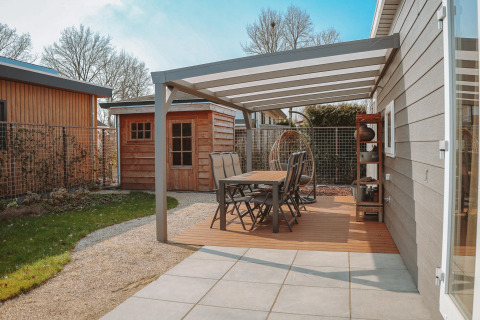 Terraza exterior con mesa de comedor y sillas bajo pérgola en Veluwelodge XL, Holiday park De Boshoek, Países Bajos.