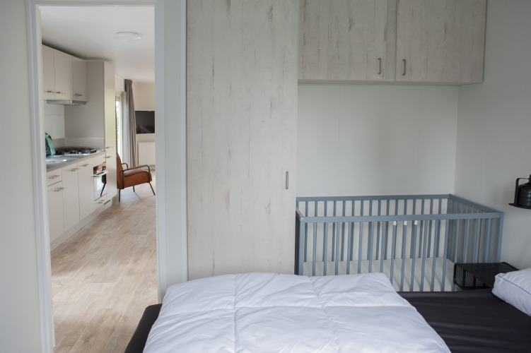 Schlafzimmer mit Babybett und Blick auf Küche und Wohnzimmer in der Veluwelodge XL, De Boshoek.