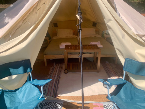 Vista interior de una tienda safari con dos sillas plegables delante y una cama doble con mesa de madera.