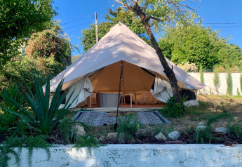 Tienda safari Oliveira Bell Standard Bell Tent con entrada abierta, rodeada de plantas verdes y naturaleza.