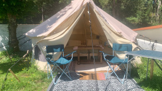 Safari-telt med åbent telt, to campingstole, et bord og tæppe på græsset under træer i solen.