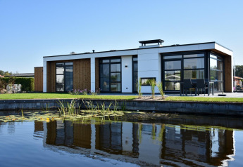 Lodge moderne avec grandes baies vitrées et bois, reflété dans un étang sous un ciel bleu clair.