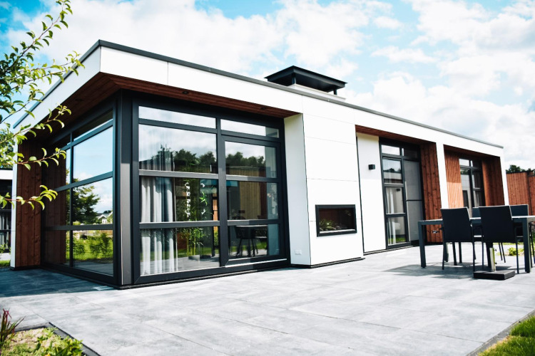 Modern lodge with large windows, outdoor seating area, surrounded by nature and a partly cloudy sky.