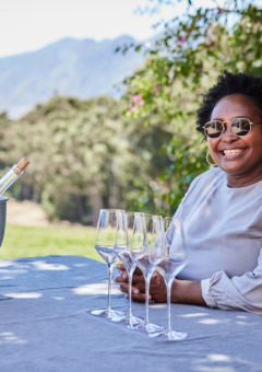 Mujer sonríe en mesa con copas de vino y hielera al aire libre en un parque de glamping pintoresco.