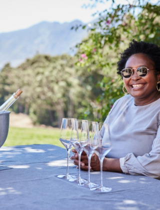 Mujer sonríe en mesa con copas de vino y hielera al aire libre en un parque de glamping pintoresco.
