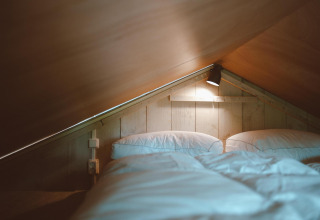 Cama en altillo con sábanas blancas y luz cálida en Tiny Safari House, Holiday park De Boshoek, Países Bajos.
