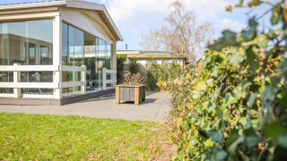 Udendørsareal ved Veluwe Lounge, en lodge med glasvægge på Holiday park De Boshoek, omgivet af hække og terrasse.