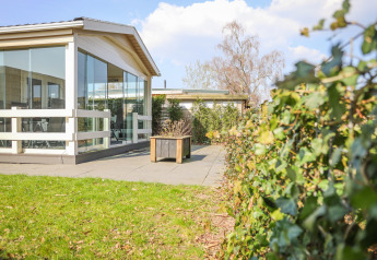 Udendørsareal ved Veluwe Lounge, en lodge med glasvægge på Holiday park De Boshoek, omgivet af hække og terrasse.