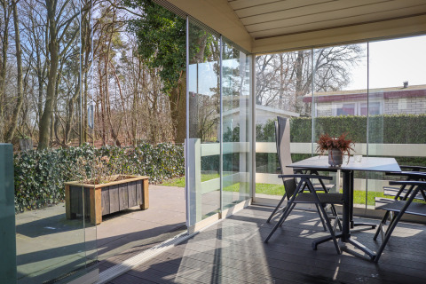 Terraza cubierta en Veluwe Lounge, Holiday park De Boshoek, Países Bajos, con paredes de vidrio y vista al bosque.