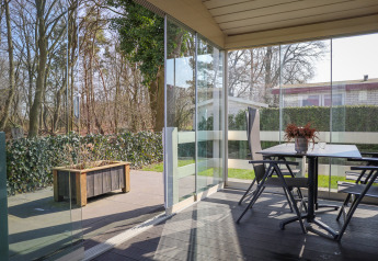 Terraza cubierta en Veluwe Lounge, Holiday park De Boshoek, Países Bajos, con paredes de vidrio y vista al bosque.