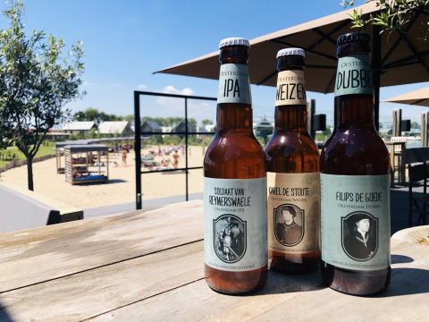 Tres botellas de cerveza artesanal sobre una mesa de madera con Villa Oesterdam y playa al fondo, Países Bajos.