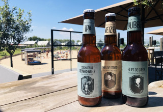 Drei lokale Bierflaschen auf Holztisch mit Oesterdam Resort, Sandstrand und Villa Oesterdam im Hintergrund.