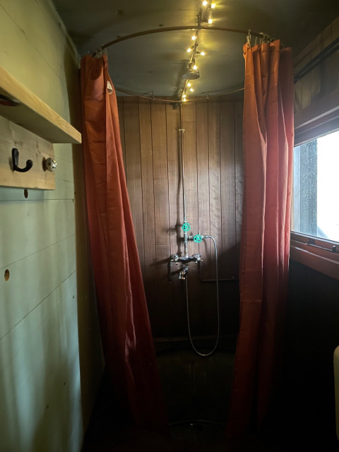 Ducha rústica con paneles de madera, cortinas naranjas y luces en el techo en un alojamiento glamping.