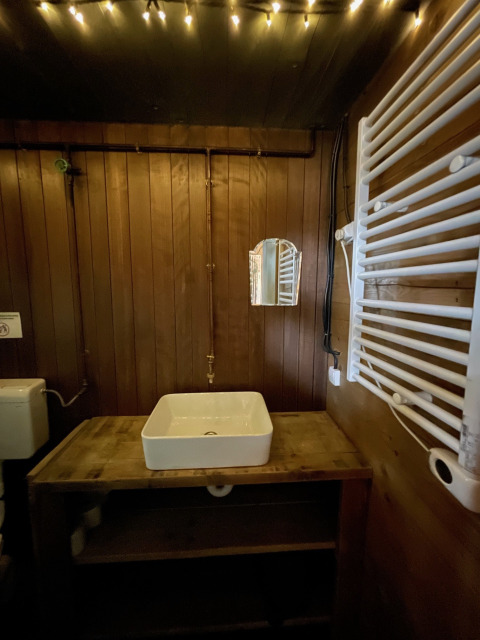 Baño de glamping con paredes de madera, lavabo cuadrado, espejo pequeño y luces de cadena cálidas.