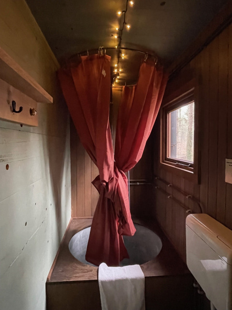 Gezellige boomhutbadkamer met een rond bad, rode douchegordijnen, sfeervolle lichtslingers en houten muren en raam.