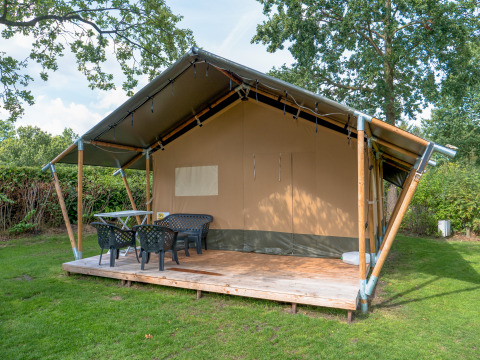 Tienda safari con plataforma de madera cubierta, sillas y mesa plásticas, rodeada de césped y árboles.