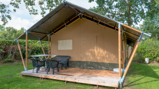 Safari tent with covered wooden deck, plastic chairs and table, set in a grassy, tree-surrounded area.