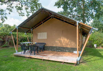 Safari tent with covered wooden deck, plastic chairs and table, set in a grassy, tree-surrounded area.