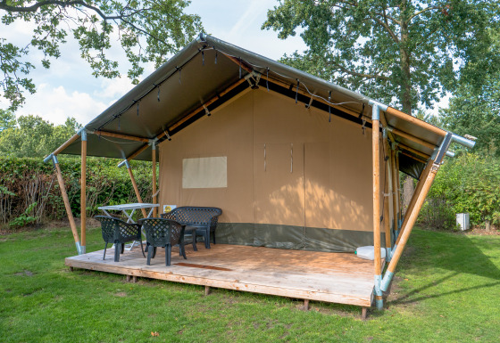 Safari tent with covered wooden deck, plastic chairs and table, set in a grassy, tree-surrounded area.