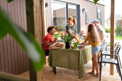 Familie isst gemeinsam auf einer Holzterrasse vor einer Lodge, sommerliche Atmosphäre im Garten.