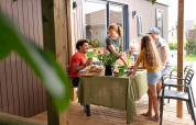 Familie nyder udendørs måltid på en træterrasse ved en lodge, omgivet af sommerstemning.