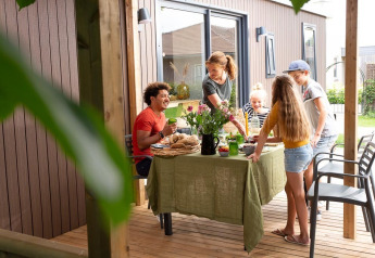 Familie isst gemeinsam auf einer Holzterrasse vor einer Lodge, sommerliche Atmosphäre im Garten.