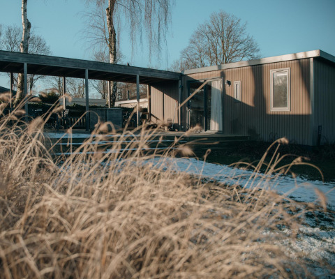 Et moderne lodgehus med træfacade og store vinduer, omgivet af tørt græs og et dækket terrasseområde.