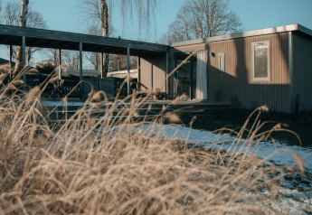 Un pavillon moderne en bois avec de grandes fenêtres, entouré d’herbes sèches et d’une terrasse ouverte.