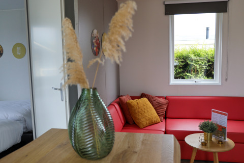 Moderne lodge stue med rødt hjørnesofa, store pyntepuder og grøn vase ved De Boshoek feriepark i Holland.