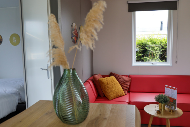 Modernes Wohnzimmer mit rotem Sofa, Kissen, grünem Glasvase und Fenster in der Family Lodge De Boshoek.