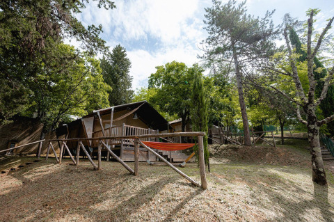 Tienda safari Villatent Wood en Camping Colleverde, Italia, rodeada de árboles y naturaleza, con hamaca delante.