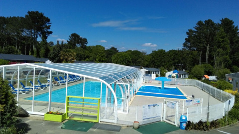 Piscina al aire libre y piscina cubierta en Camping de Kerleyou, rodeadas de vegetación en Bretaña, Francia.