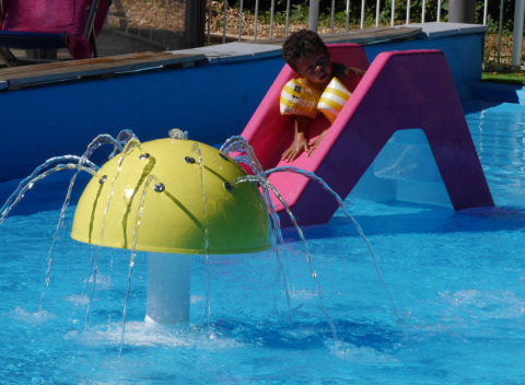 Niño con flotadores bajando por un tobogán rosa junto a una fuente amarilla en una piscina infantil.
