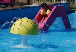 Bambino con braccioli scivola su uno scivolo rosa vicino a una fontana gialla in piscina all'aperto.