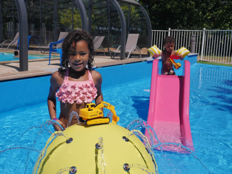Niños juegan en la piscina con excavadoras de juguete, un tobogán rosa y chorros de agua en Camping lac Aux Oiseaux.