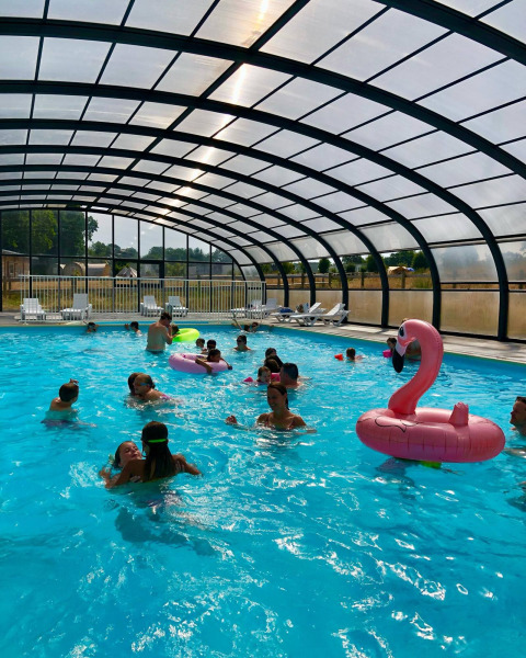 Piscina cubierta con niños, adultos y un flotador de flamenco rosa en Camping O2 en Normandía, Francia.