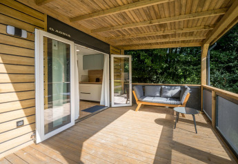 Terraza de madera cubierta en Lodge at Urban-Gardens Ieper, Bélgica, con sofá, mesa y vistas verdes.
