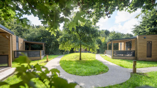 Modern wooden lodges encircle a grassy lawn with winding paths, surrounded by lush trees and nature.