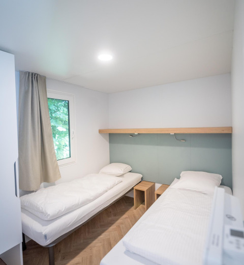 Chambre de lodge lumineuse avec deux lits simples, linge blanc, fenêtre et mobilier en bois clair.