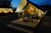 Beleuchtetes Safari-Zelt mit Holzterrasse und Sitzmöbeln bei Urban-Gardens Ieper, Belgien, abends.