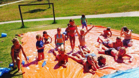 Children play and slide on a large orange tarp with water in the sun at a holiday park in Marche, Italy.