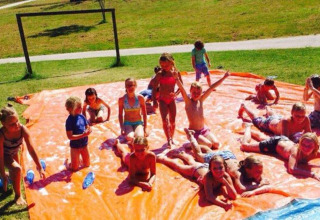 Kinder spielen fröhlich auf einer großen orangenen Plane mit Wasser bei einem Ferienpark in Marche, Italien.