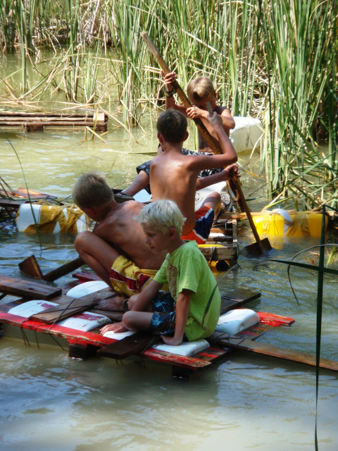 Bambini su una zattera fatta in casa tra le canne all'acqua presso Camping Podere sei Poorte, Marche, Italia.