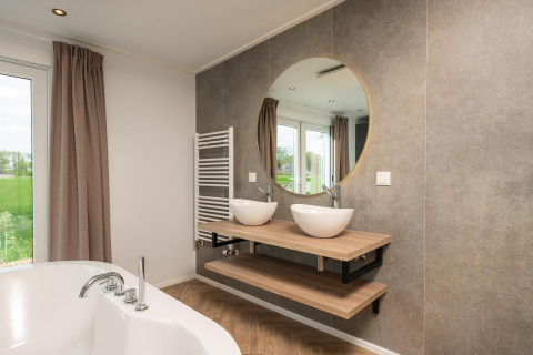 Baño moderno con lavabos dobles, espejo redondo y vista exterior en Panorama Lodge, Vakantiepark Hölte.