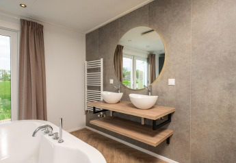 Baño moderno con lavabos dobles, espejo redondo y vista exterior en Panorama Lodge, Vakantiepark Hölte.