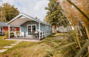 Chalet lumineux avec terrasse, mobilier de jardin et pelouse, entouré d’arbres sous un ciel ensoleillé.