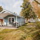 Helle Ferienlodge mit Terrasse, Gartenmöbeln und Rasen, umgeben von Bäumen, bei sonnigem Wetter.