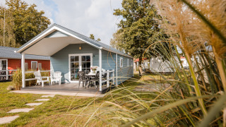 Zonnige lodge met veranda en tuinmeubilair, te midden van groen, gras en bomen onder een blauwe hemel.