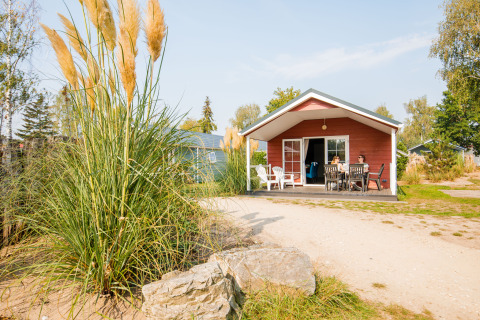 Buitenfoto van een knus rood verblijf met veranda, waar twee mensen aan een tafel zitten in de zon.